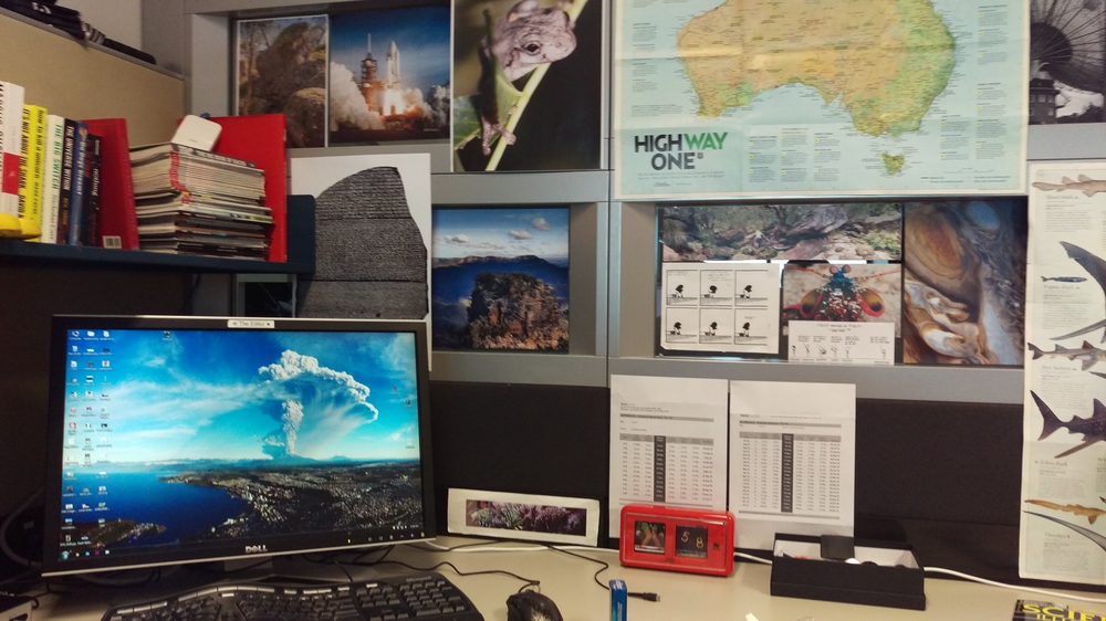 An office desk under a wall of science posters: an Australia map, a space shuttle launch, a tree frog, the Rosetta Stone and sharks